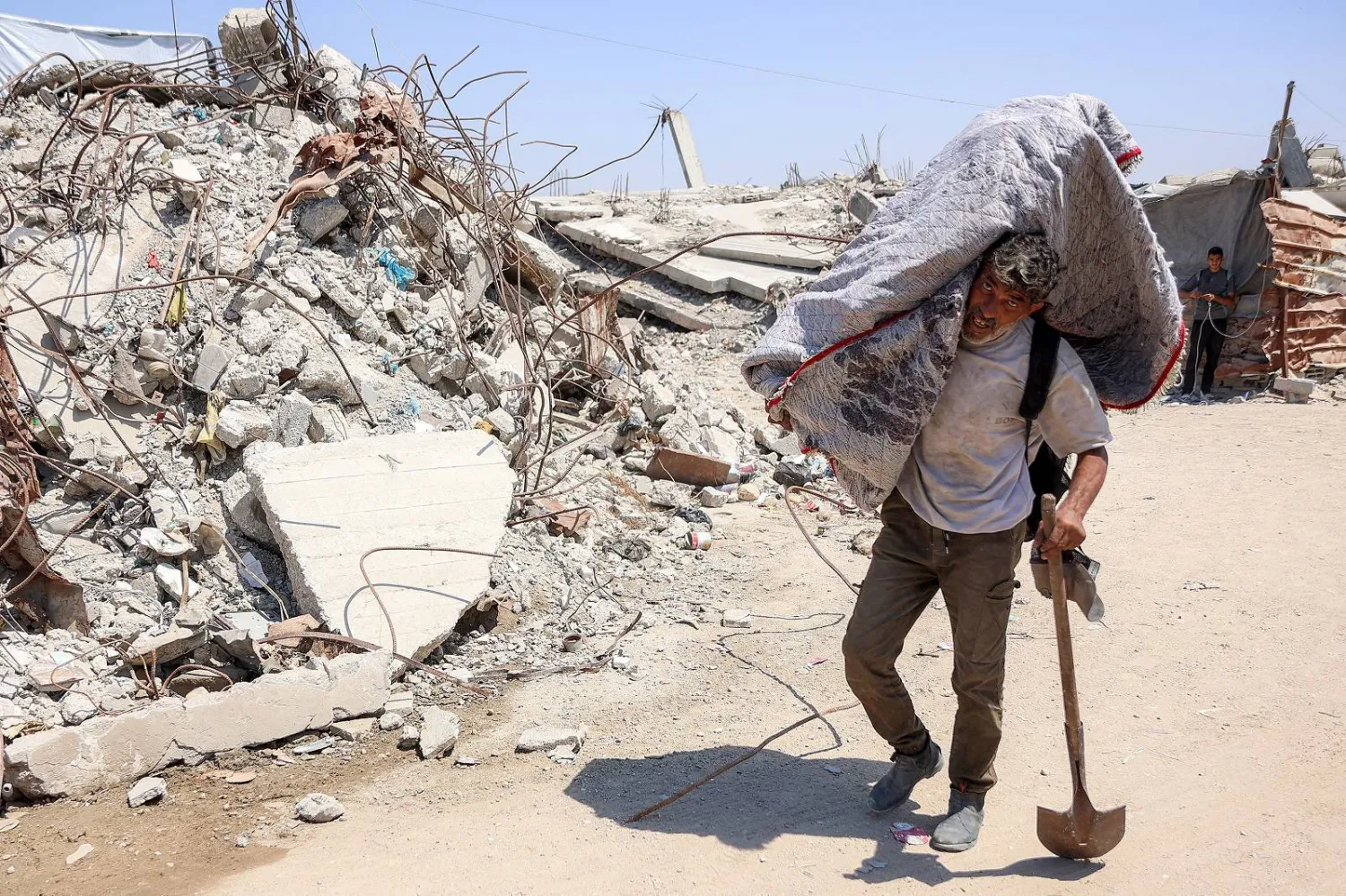 فلسطيني يحمل أمتعة على كتفه ويمر بمحاذاة الأنقاض في مخيم للنازحين بغزة (أ.ف.ب)
