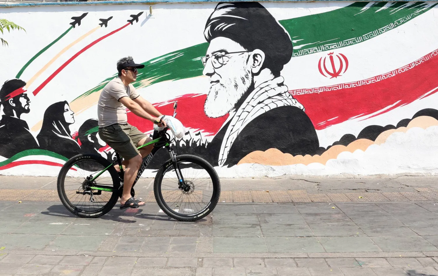 An Iranian man passes with his bike next to a wall painting featuring slain Iranian supreme leader Ali Khamenei in a street in central Tehran, Iran, 13 August 2026. (EPA)