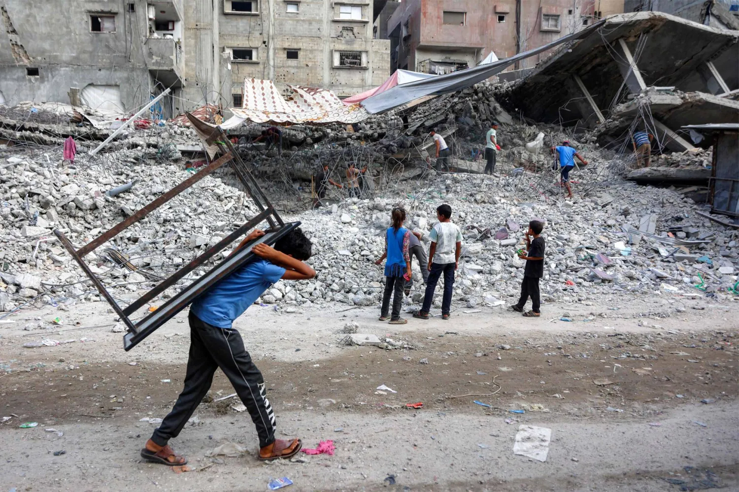 صبي فلسطيني يحمل طاولة يمر قرب عمال يجمعون أنقاض مبان دمرتها إسرائيل في الحرب على غزة (أ.ف.ب)