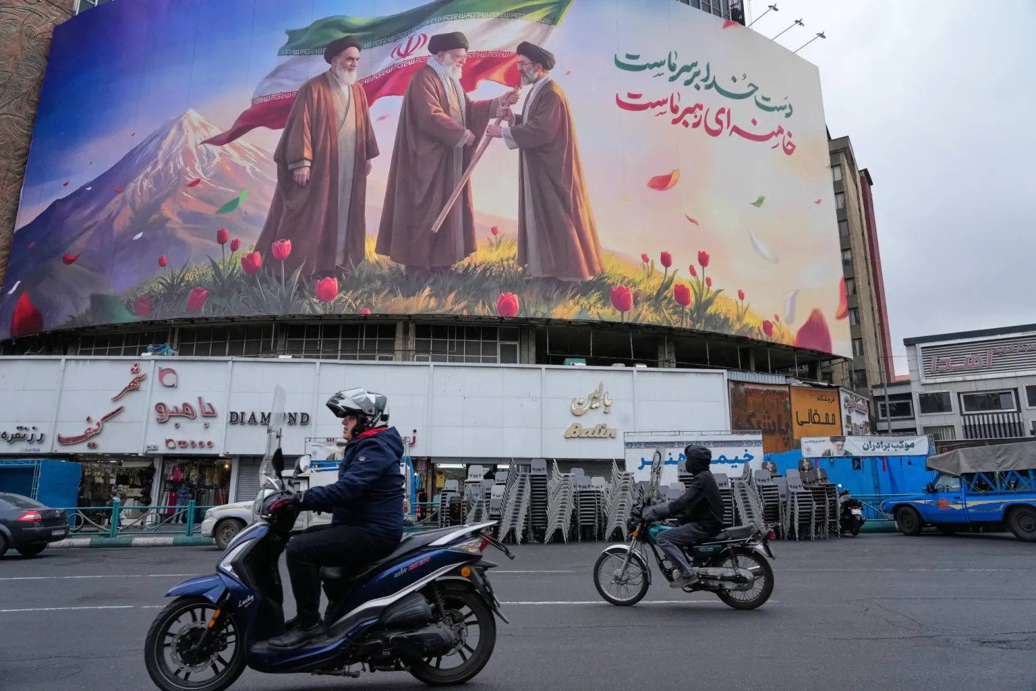 Motorbikes drive past a billboard depicting Iran's late Supreme Leader Ali Khamenei, center, handing the country's flag to his son and successor, Mojtaba Khamenei, right, as the late revolutionary founder Khomeini stands at left, in a square in downtown Tehran, Iran, March 10, 2026. (AP) 