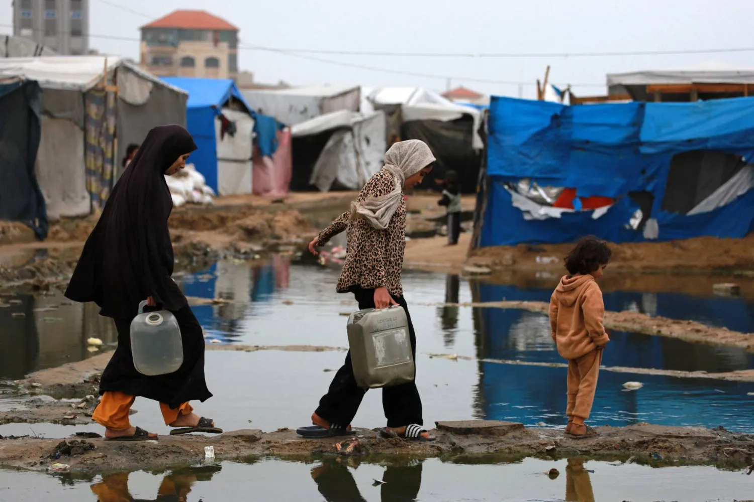 تستخدم نساء فلسطينيات نازحات طريقاً جافاً لنقل المياه إلى خيمتهن بعد هطول أمطار غزيرة شمال دير البلح في وسط قطاع غزة (أ.ف.ب)