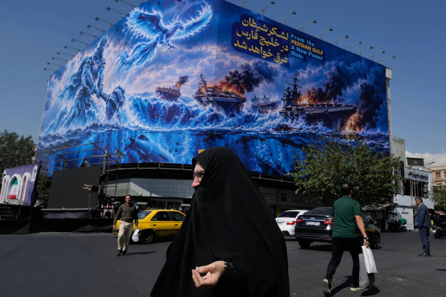 Pedestrian walk past a billboard depicting a Persian legendary hero and flying figure hitting US aircraft carriers alongside the message in English and Farsi: "From the Gulf a new power rises" "The army of Satan will be drowned in the Gulf, " at revolution square in downtown Tehran, Iran, Monday, Sept. 14, 2026. (AP) 