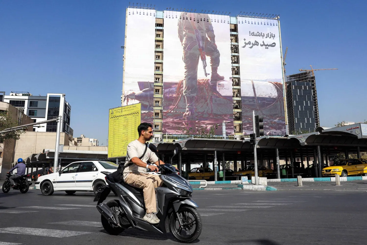 A motorist rides a scooter near a giant billboard erected on the facade of a building depicting a member of Iran's Revolutionary Guards Corps (IRGC) placing his foot on a US sea drone, reportedly seized by Iran in the Strait of Hormuz, in Vanak Square in Tehran on September 24, 2026. (Photo by AFP) 