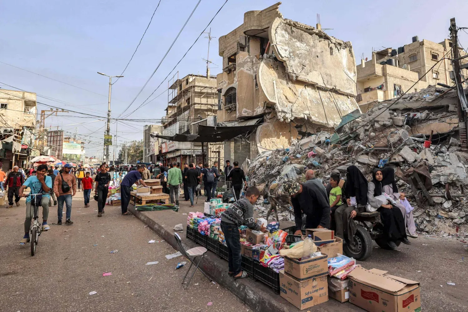 Nisan 2024’te Gazze Şeridi’nin güneyindeki Refah’ta yıkılmış bir binanın enkazı önünde duran bir satıcıya ürünlerini düzenlemesi için yardım eden çocuk (AFP)
