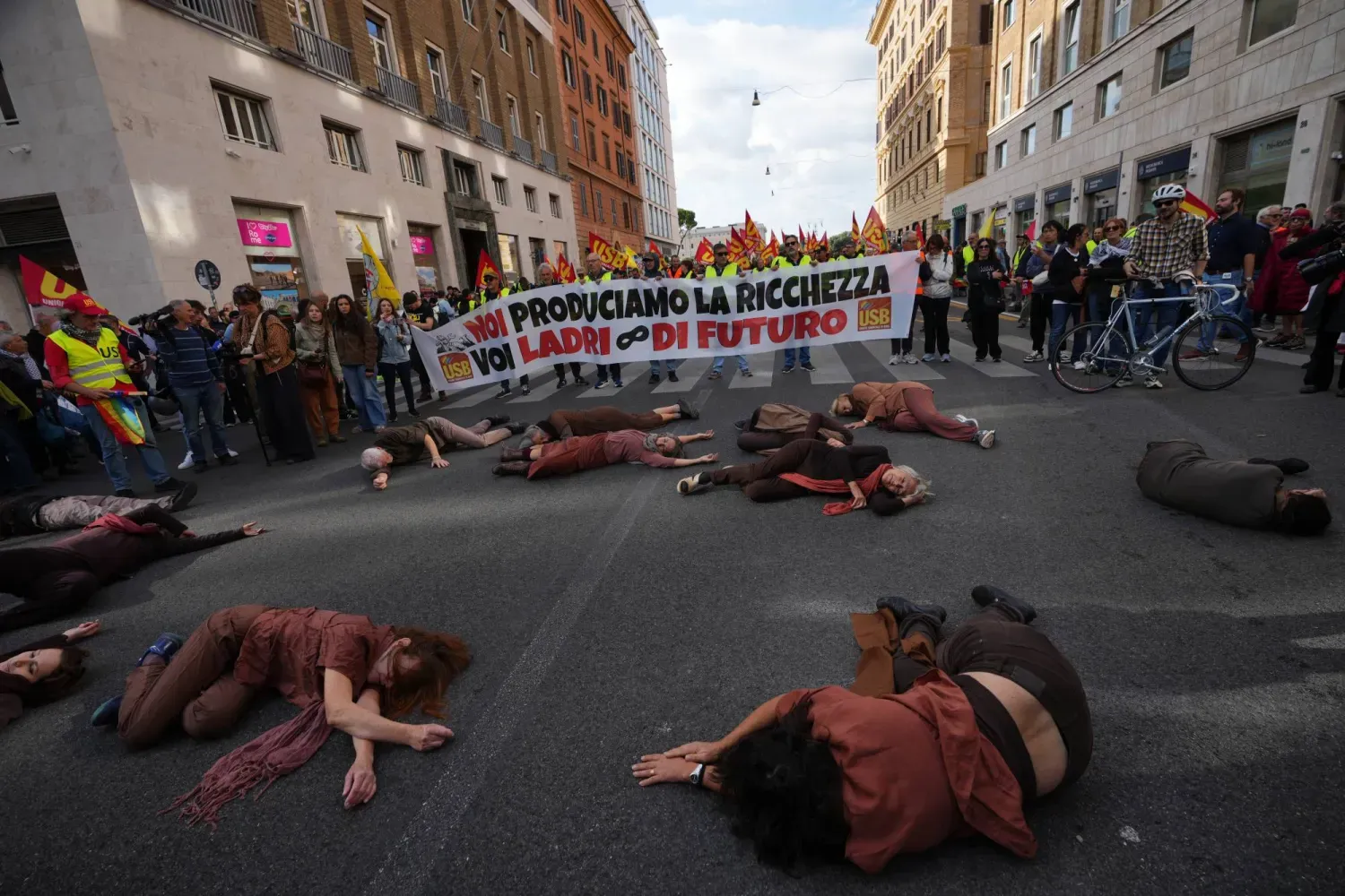 İtalya’nın başkenti Roma’da, hayat pahalılığı, düşük ücretler ve askeri harcamaları protesto etmek amacıyla düzenlenen genel grev sırasında, üzerinde “Serveti biz üretiyoruz, siz ise geleceğimizi çalıyorsunuz” yazılı pankart taşıyan göstericilerin önünde yere uzanan performans sanatçıları, 30 Ekim 2026 (AP)