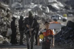 Gazze Şeridi'nin kuzeyindeki Cibaliye Mülteci Kampı’nda İsrailli rehinelerin cesetlerini arayan İzzeddin el-Kassam Tugayları savaşçılarının yanında duran Filistinli bir çocuk, 1 Aralık 2025 (EPA)