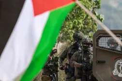 A Palestinian flag flutters in front of Israeli soldiers standing near their military vehicle parked at the entrance of the Nur Shams Palestinian refugee camp, in the Israeli-occupied northern West Bank on December 15, 2025. (AFP) 