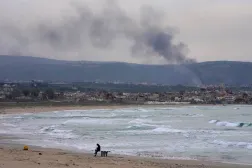 A man checks his phone on a beach as smoke rises from Israeli artillery shells on Qlaileh village, visible from Tyre city, south Lebanon, March 28, 2026. (AP)