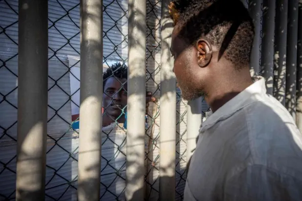 مهاجر يتحدث مع رجل عبر جدار في مركز مؤقت لطالبي اللجوء بمدينة مليلية (أ.ف.ب)