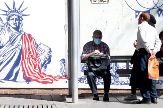 انتظار تعدادی از افراد در ایستگاه اتوبوسی مقابل نقاشی‌های دیواری در روی دیوار سفارت سابق آمریکا در تهران که میز مذاکره را به تصویر کشیده‌اند (EPA)