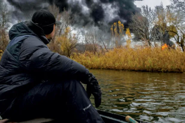 صياد يبحر بقاربه على نهر دنيبرو بينما تتصاعد ألسنة دخان من احتياطي نفطي في خيرسون أمس (أ.ف.ب)