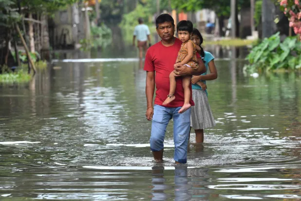 رجل يحمل طفلة ويسير وسط شارع غمرته المياه بعد هطول الأمطار الموسمية في باتسالا بولاية آسام الهندية (أ.ف.ب)
