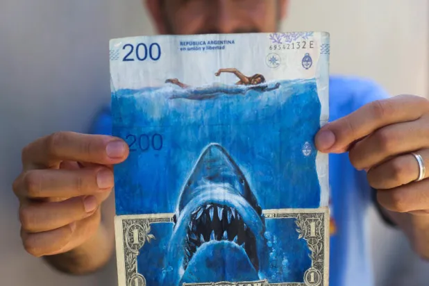 دياز يحمل أوراق بيزو متداخلة مع الدولار تصور فيلم ستيفن سبيلبرغ «القرش» باعتباره محاكاة ساخرة للتضخم المتزايد في الأرجنتين (رويترز)