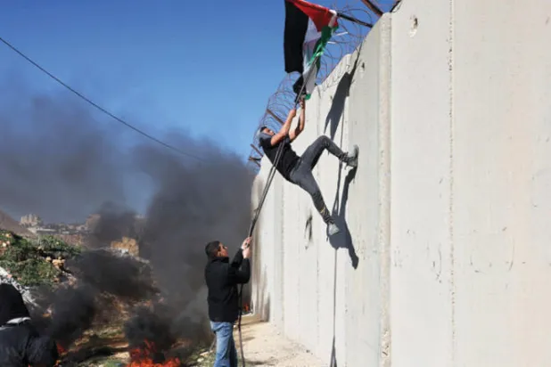 فلسطينيون يرفعون علماً فوق الجدار الفاصل الإسرائيلي بالضفة الجمعة احتجاجاً على توسيع مستوطنة عطاروت (أ.ف.ب)
