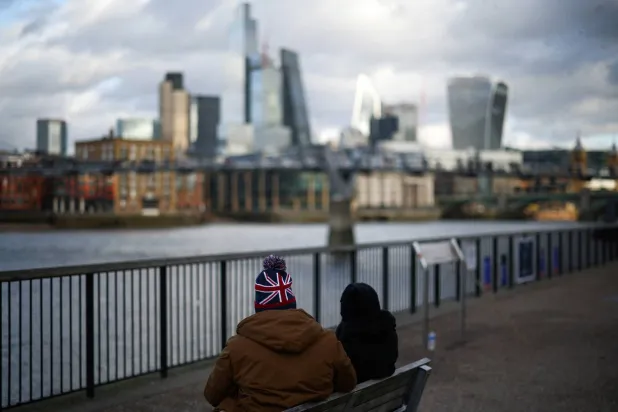 أشخاص يجلسون على ضفة نهر التيمس مع ظهور الحي المالي لمدينة لندن في الخلفية ببريطانيا (رويترز)