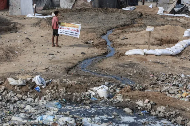 طفل فلسطيني يحمل لافتة مكتوباً عليها «ممنوع الدخول... مشروع الصرف الصحي» التي وُضعت بالقرب من أكوام القمامة التي أُلقيت بين خيام النازحين في دير البلح (د.ب.أ)