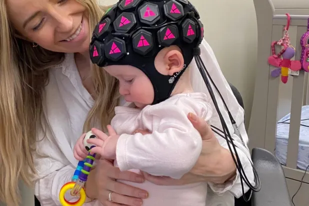 صورة لإحدى الأمهات وطفلها ممن تم إجراء التجربة عليهم ويظهر فيها الجهاز على رأس الطفل ويتيح له حرية الحركة ما يسمح برصد النشاط العصبي (كلية بيركبيك)