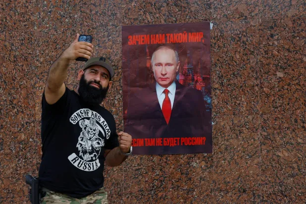 رجل يلتقط صورة تذكارية مع ملصق يحمل صورة الرئيس الروسي فلاديمير بوتين يقول: «لماذا نريد مثل هذا العالم إذا لم تكن روسيا موجودة فيه؟» (رويترز)
