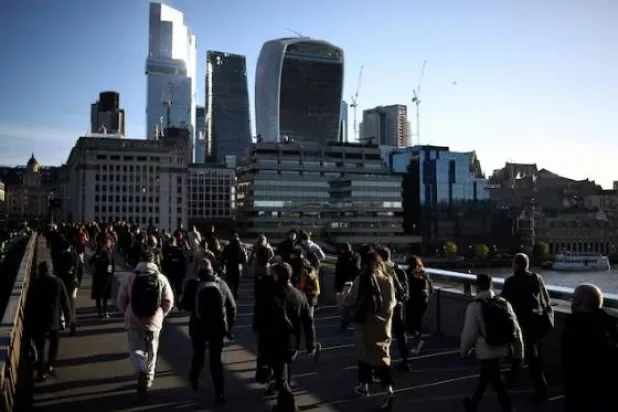 يسير الناس عبر جسر لندن خلال ساعة الذروة الصباحية مع الحي المالي للمدينة في الخلفية (رويترز)