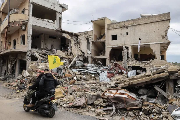 رجال يركبون دراجة نارية تحمل علم «حزب الله» ويمرون بجوار مبانٍ مدمرة بينما يتجه الناس إلى منازلهم في جنوب لبنان (أ.ف.ب)