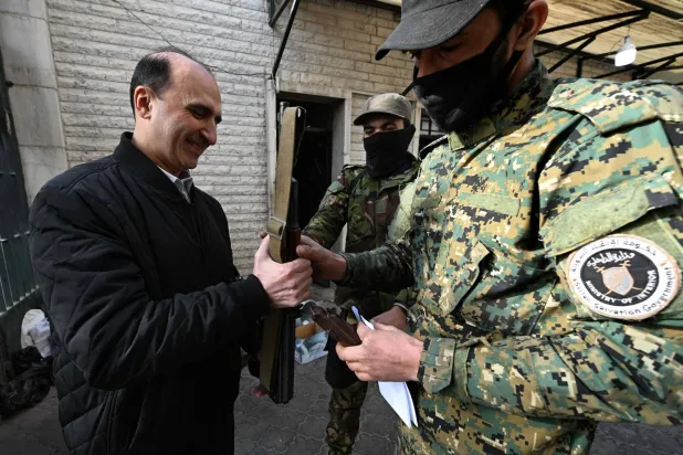 جندي من «حكومة الإنقاذ» التابعة لـ«هيئة تحرير الشام» (يمين) يتسلم سلاح أحد حراس الأمن السابقين بمكاتب حزب البعث بدمشق 12 ديسمبر 2024 (أ.ف.ب)