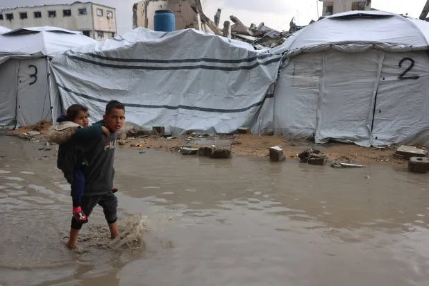 طفل فلسطيني نازح يحمل أخته أثناء عبوره بركة مياه بالقرب من مخيم مؤقت خلال عاصفة في مدينة غزة (أ.ف.ب)