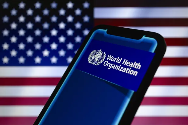 شعار منظمة الصحة العالمية يظهر على هاتف ذكي وخلفه علم الولايات المتحدة (د.ب.أ)