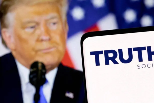 شعار شبكة «تروث سوشيال» على هاتف ذكي أمام شاشة تظهر الرئيس الأميركي دونالد ترمب (رويترز)