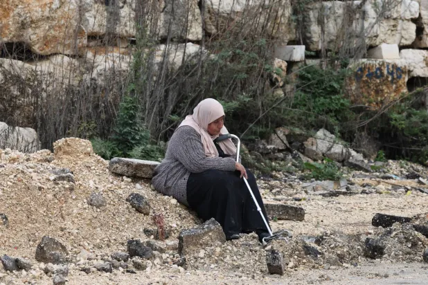 امرأة فلسطينية مسنة تُمسك بعكازها وتستريح على جانب طريق متضرر في مخيم نور شمس للاجئين بالقرب من مدينة طولكرم بالضفة الغربية (إ.ب.أ)