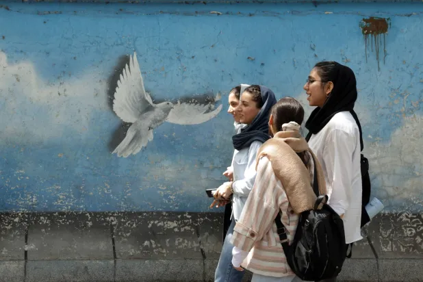 فتيات إيرانيات يَسِرْن أمام لوحة جدارية بأحد شوارع طهران (إ.ب.أ)