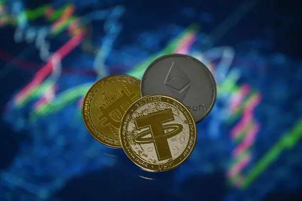 عملات مشفّرة تذكارية من «تيذر» و«بتكوين» و«إيثيريوم» بجانب شاشة تداول (أ.ف.ب) 