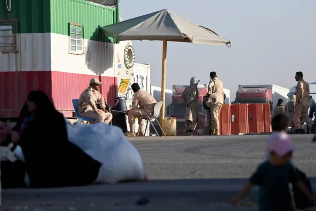 يقف ضباط الحدود الإيرانيون حراساً بينما يصل اللاجئون الأفغان إلى نقطة الصفر في معبر إسلام قلعة الحدودي بين أفغانستان وإيران في 28 يونيو الحالي بعد ترحيلهم من إيران (أ.ف.ب)