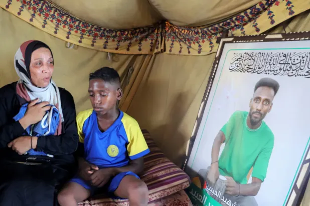 Doaa, the widow of late soccer player Suleiman Al-Obeid, known as the "Palestinian Pele," who was killed by an Israeli strike targeting people waiting for humanitarian aid, according to the Palestine Football Association, looks at his picture while holding his shorts, as her son sits beside her inside their tent in Gaza City August 9, 2025. REUTERS/Ebrahim Hajjaj 