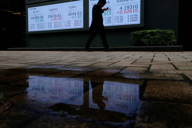 رجل يمر أمام لوحة أسعار أسهم تُظهر مؤشر داو جونز ومتوسط ​​أسهم «نيكي» وعائد سندات الحكومة اليابانية والأميركية في طوكيو (رويترز)