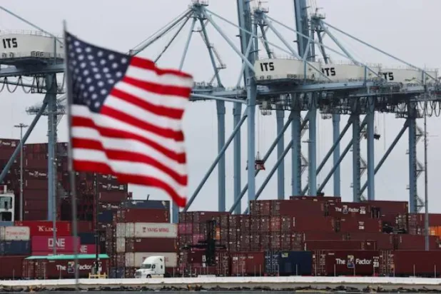 علم الولايات المتحدة يرفرف أمام حاويات شحن في ميناء لونغ بيتش بولاية كاليفورنيا (رويترز)
