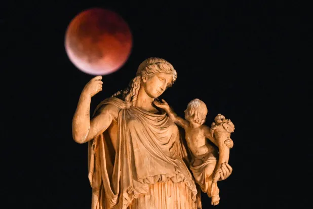 «القمر الدموي» أثناء خسوف القمر مع تمثال للإلهة اليونانية القديمة إيرين تحمل طفلاً في المقدمة (أ.ف.ب)