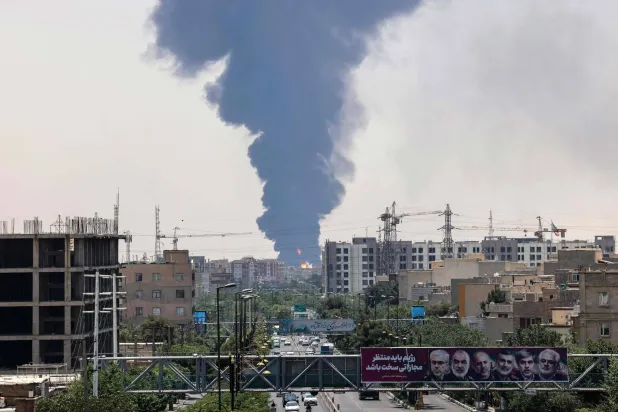 صور الجنرالات والعلماء النوويين الإيرانيين الذين قُتلوا خلال الغارات الإسرائيلية معلقة في شارع بطهران ووراءها دخان يتصاعد من موقع استُهدف 15 يونيو الماضي (أ.ف.ب)