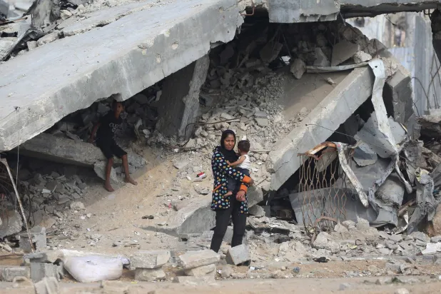 فلسطينية تحمل طفلها بين مبانٍ مدمرة على طريق قرب مخيم النصيرات للاجئين في وسط قطاع غزة (أ.ف.ب)