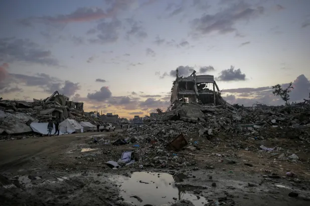 فلسطينيون يسيرون بالقرب من مبانٍ مدمرة في يوم ممطر شرق مدينة غزة - 16 نوفمبر 2025 (إ.ب.أ)