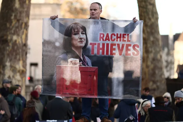 متظاهر يرفع لافتة كُتب عليها راشيل ثيفز مقابل داونينغ ستريت في اليوم الذي قدمت فيه ريفز بيان الموازنة (رويترز)