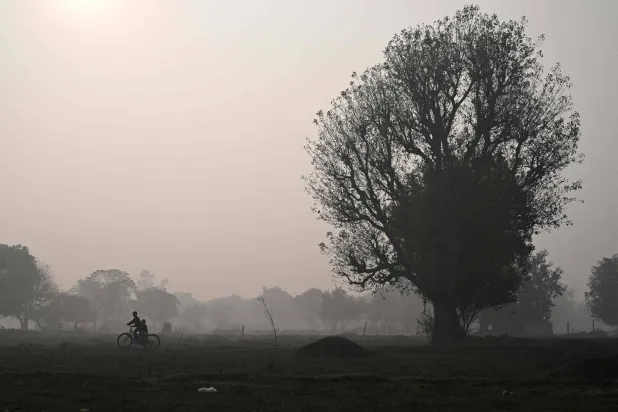 Children ride a bicycle across a field on smoggy winter morning in New Delhi on December 17, 2025. (Photo by Arun SANKAR / AFP)