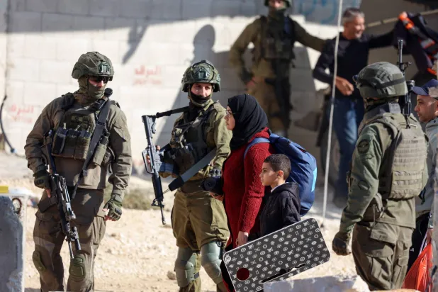 A Palestinian resident of the Nur Shams refugee camp walks with a child past Israeli soldiers while carrying belongings retrieved from her home ahead of the Israeli military's demolition of residential buildings in the camp near Tulkarem in the Israeli-occupied West Bank on December 17, 2025. (AFP)