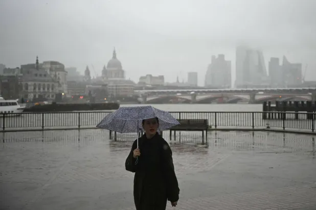 A pedestrian shelters from the rain while walking along the South Bank with the River Thames and the office buildings of the City Of London shrouded in fog on December 18, 2025. (Photo by JUSTIN TALLIS / AFP)