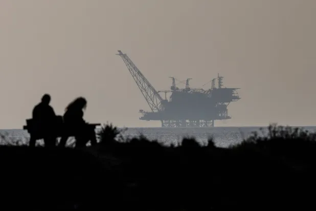 شخصان يجلسان على مقعد وخلفهم حقل غاز ليفياثان في محمية شاطئ هابونيم الطبيعية شمال إسرائيل (إ.ب.أ)