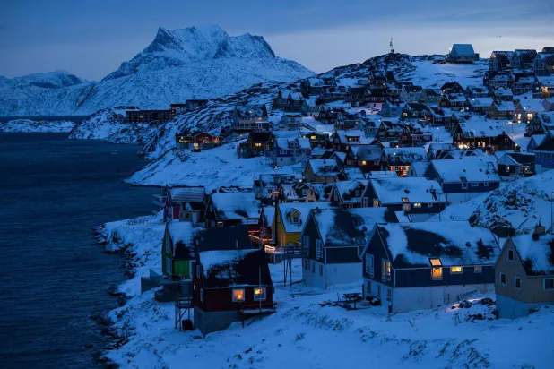 منازل مضاءة على طول الساحل مع بزوغ ضوء الصباح الباكر فوق التلال المغطاة بالثلوج في نوك بغرينلاند 22 يناير 2026 (أ.ف.ب)