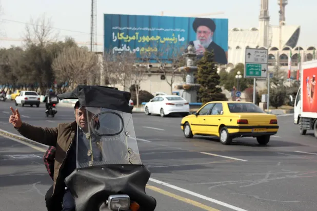 رجل إيراني يرفع علامة النصر أثناء مروره بجوار صورة للمرشد علي خامنئي في أحد شوارع طهران (إ.ب.أ)