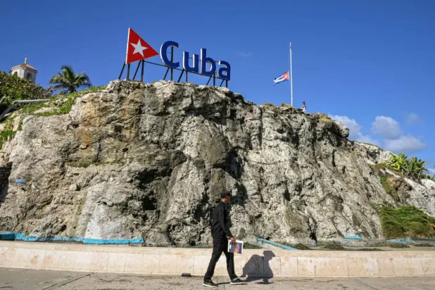 Havana'da Kübalı bir adam, üzerinde Küba yazılı bir tabelanın ve yarıya indirilmiş ulusal bayrağın önünden geçiyor (AFP)