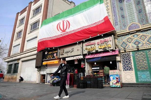 An Iranian man speaks on his mobile phone as he walks past a huge national flag hanging above shops, in Tehran on February 6, 2026. (AFP)
