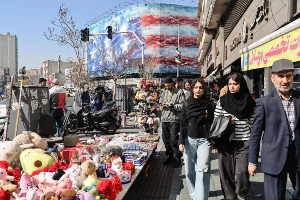 إيرانيون يمرون أمام سلع يعرضها بائع متجول مع لافتة معادية للولايات المتحدة تظهر في الخلفية بساحة انقلاب (الثورة) وسط طهران (أ.ف.ب)