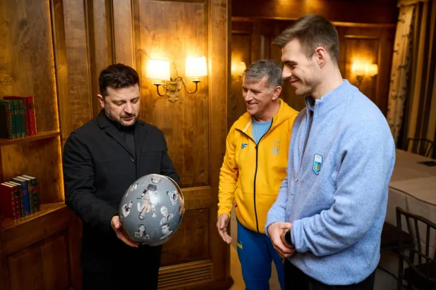 Ukrainian President Volodymyr Zelenskiy holds helmet as he meets with a Ukrainian skeleton racer Vladyslav Heraskevych , who was disqualified from the Olympic skeleton competition over his "helmet of remembrance" depicting athletes killed since Russia's invasion and his father and coach, Mykhailo Heraskevych, amid Russia's attack on Ukraine, in Munich, Germany February 13, 2026. (Ukrainian Presidential Press Service/Handout via Reuters) 